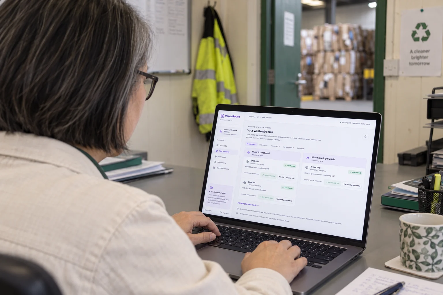 AI illustration viewed over a supplier administrator’s shoulder as she uses the PaperRoute supplier services screen on a laptop